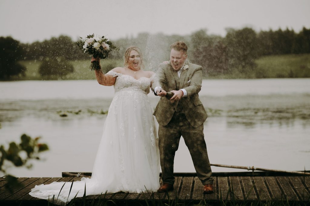 A bride and groom spray a bottle of champagne in front of a lake for their couples portraits.